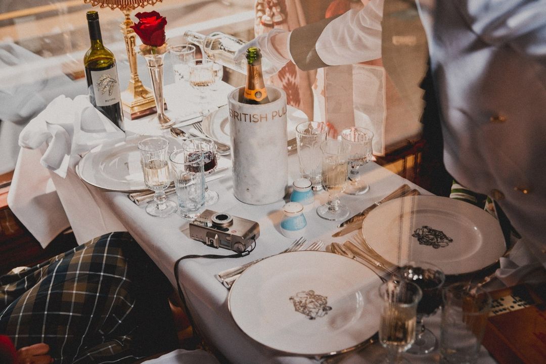 Elegant dining table set with wine, champagne, glassware, and silverware, prepared by a server with a vintage camera nearby.