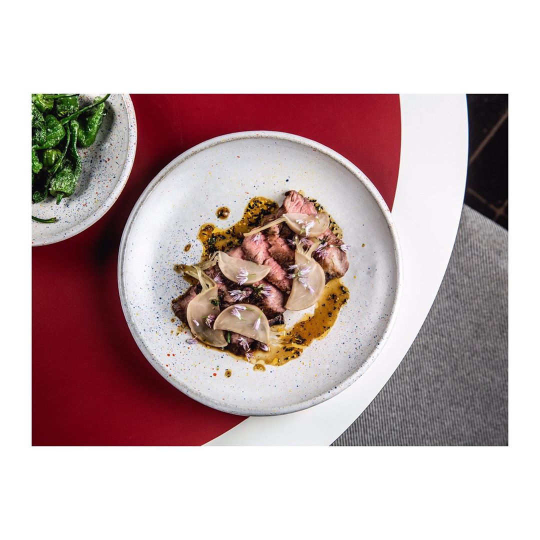 Overhead shot of a plate with sliced meat, pale turnips, and sauce, plus a side plate of padron peppers on a red and white table.