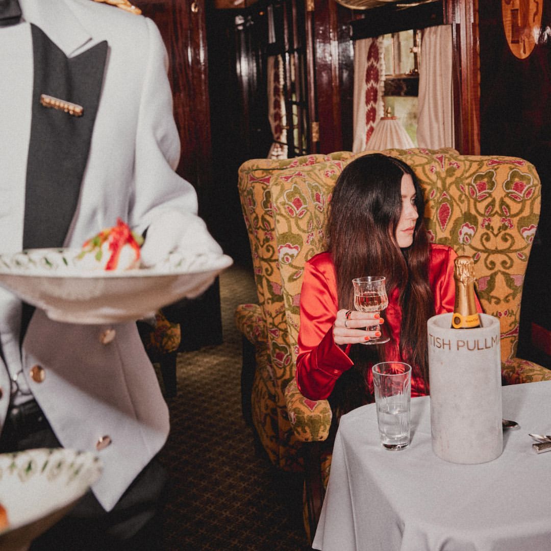 Elegant woman in red silk sits in a vintage train car. A waiter serves a dish of food. Bottle of champagne on the table.