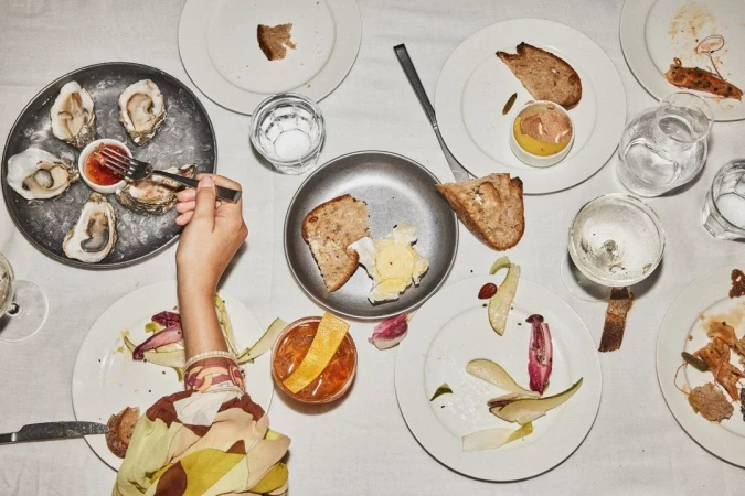 Overhead shot of a table featuring oysters on ice, plates with food remnants, bread, and an Old Fashioned cocktail. A person is using a fork to eat an oyster.
