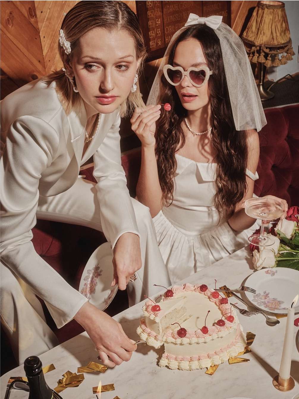 Two women in white dresses celebrate with a cake, champagne, and candles at a table, suggesting a wedding or special event in an indoor setting.