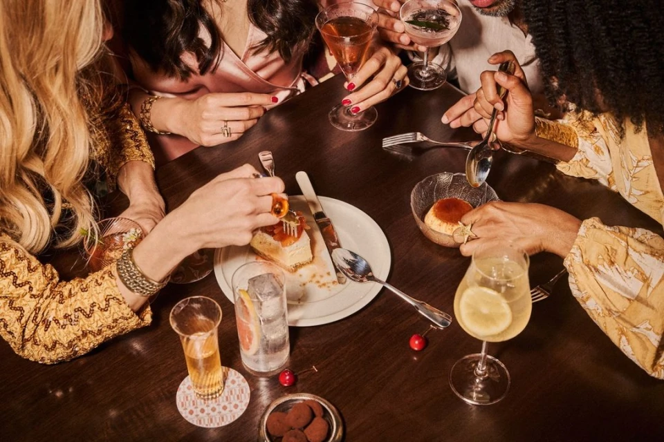 Overhead shot of friends sharing desserts and drinks at a dark wooden table with cocktails, cake, truffles, and spoons.
