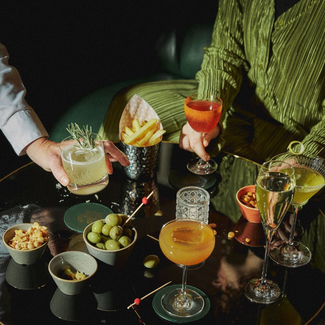 Elevated view of a cocktail table setting with mixed drinks, olives, and snacks. Two people in the background hold drinks.
