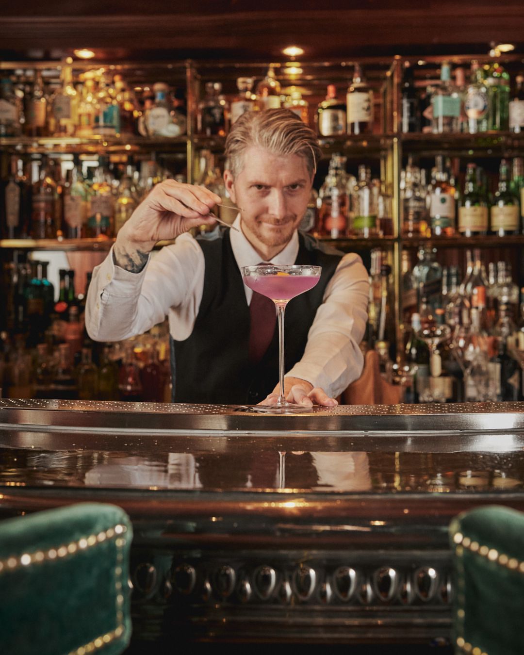 Bartender in a bar preparing a purple cocktail using a bar spoon, with an assortment of bottles on the bar in the background.