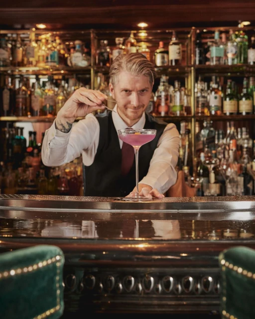Bartender in a bar preparing a purple cocktail using a bar spoon, with an assortment of bottles on the bar in the background.