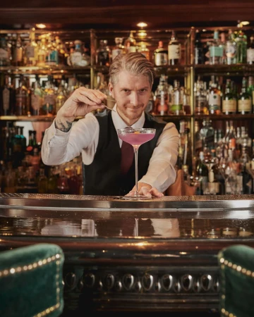 Bartender in a bar preparing a purple cocktail using a bar spoon, with an assortment of bottles on the bar in the background.