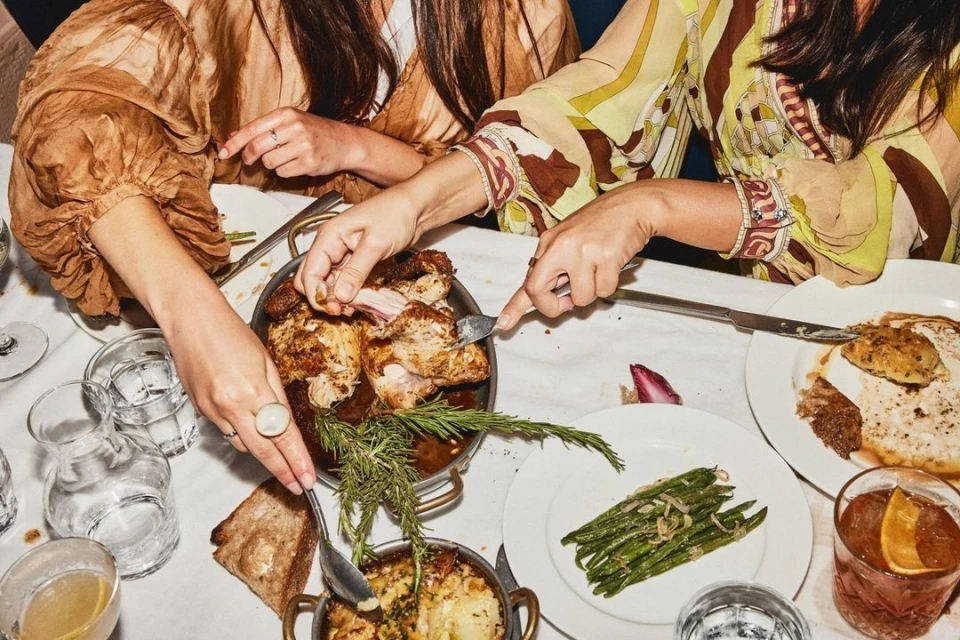 Overhead shot of three people sharing a meal of roast chicken, potatoes au gratin, and green beans on a white tablecloth-covered table.