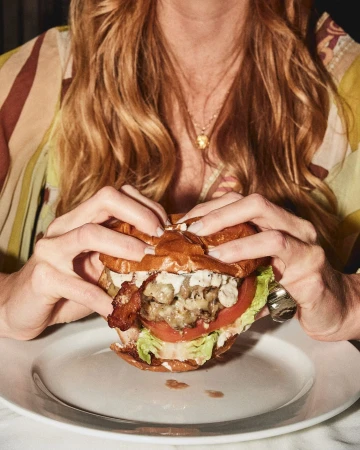 Close-up of a woman holding a burger topped with bacon, tomato, lettuce, and creamy sauce, displayed on a white plate.