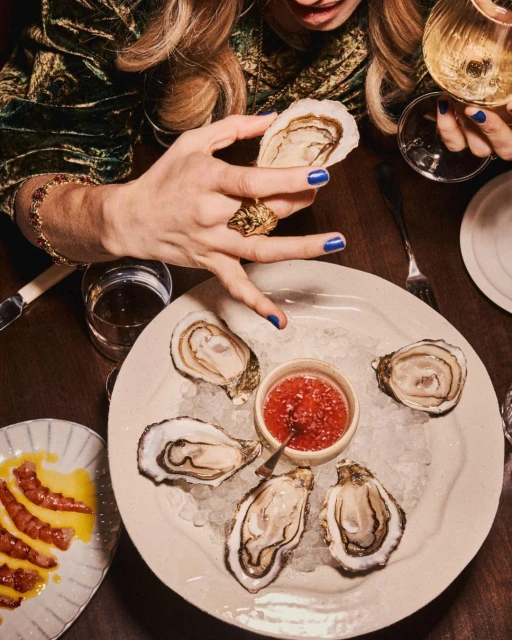 Overhead shot of a woman holding an oyster, plate of oysters on ice with cocktail sauce, and drinks on a dark wooden table.