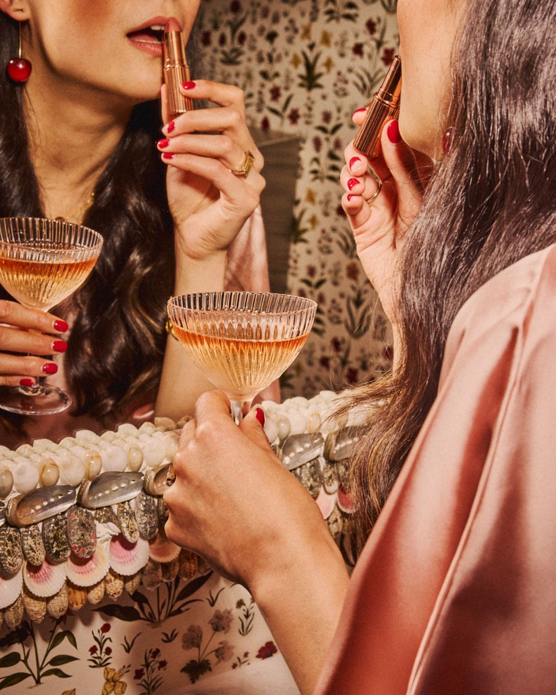 Woman in a pink silk robe applying lipstick, reflected in a vintage mirror with a champagne glass, against a floral wallpaper backdrop.