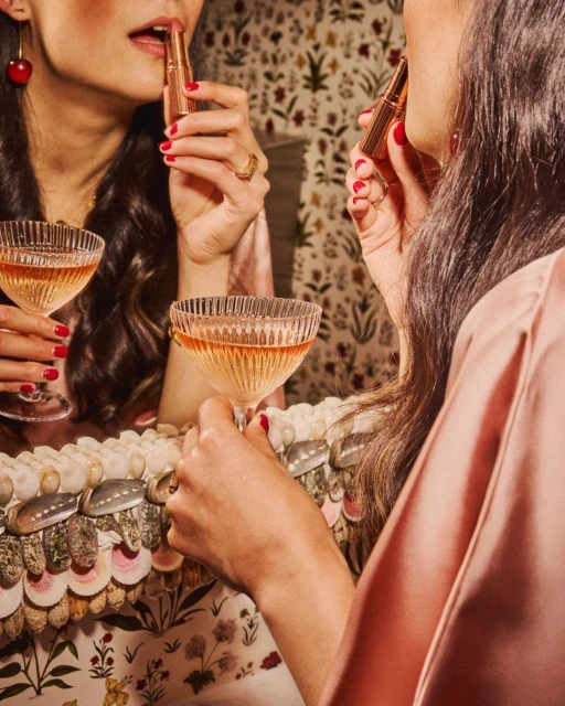 Woman in a pink silk robe applying lipstick, reflected in a vintage mirror with a champagne glass, against a floral wallpaper backdrop.
