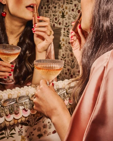 Woman in a pink silk robe applying lipstick, reflected in a vintage mirror with a champagne glass, against a floral wallpaper backdrop.
