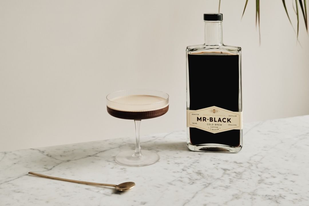 Espresso martini drink in coupe glass next to Mr. Black coffee liqueur bottle and gold spoon on marble surface, indoors.