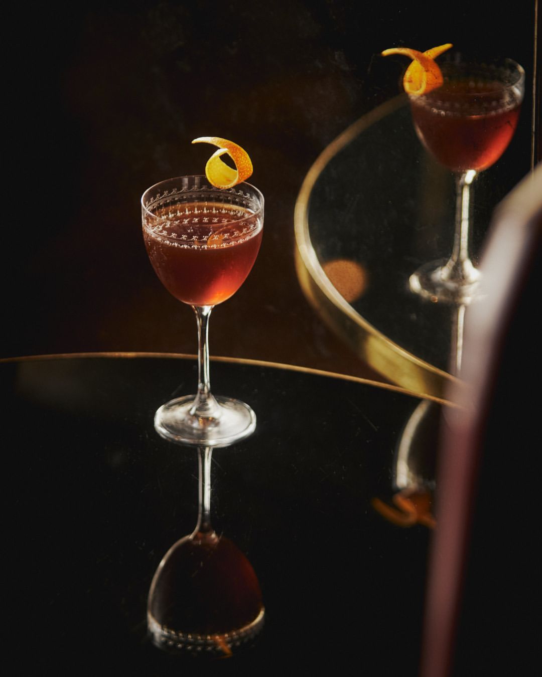 Elegant cocktail in stemmed glass on a reflective black surface, garnished with an orange peel, mirrored with warm, muted tones.