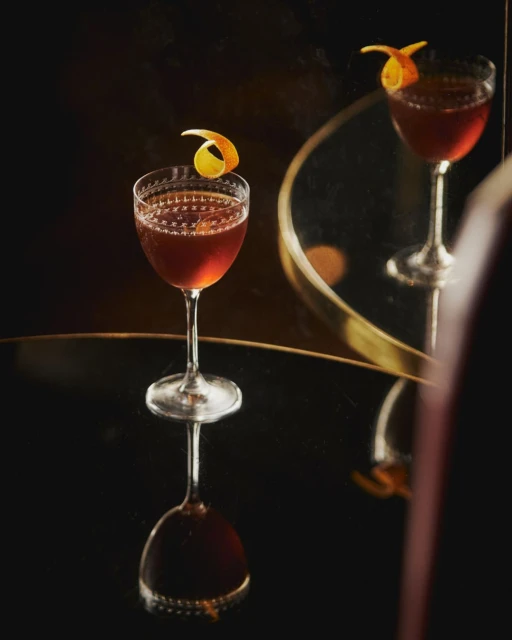 Elegant cocktail in stemmed glass on a reflective black surface, garnished with an orange peel, mirrored with warm, muted tones.