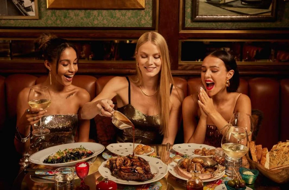 Three women dine together in a warmly lit restaurant. Food is served on plates on a table. One woman pours sauce over food.