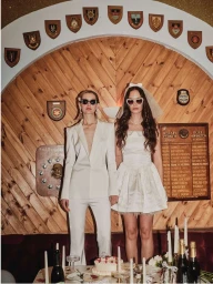 Two women wearing white outfits and sunglasses standing in front of a wooden paneled wall with crests by Charlie Mckay