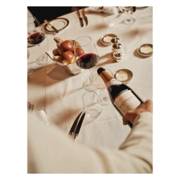 A person pouring wine from a bottle at a elegantly set dining table with bread rolls and glassware in London.