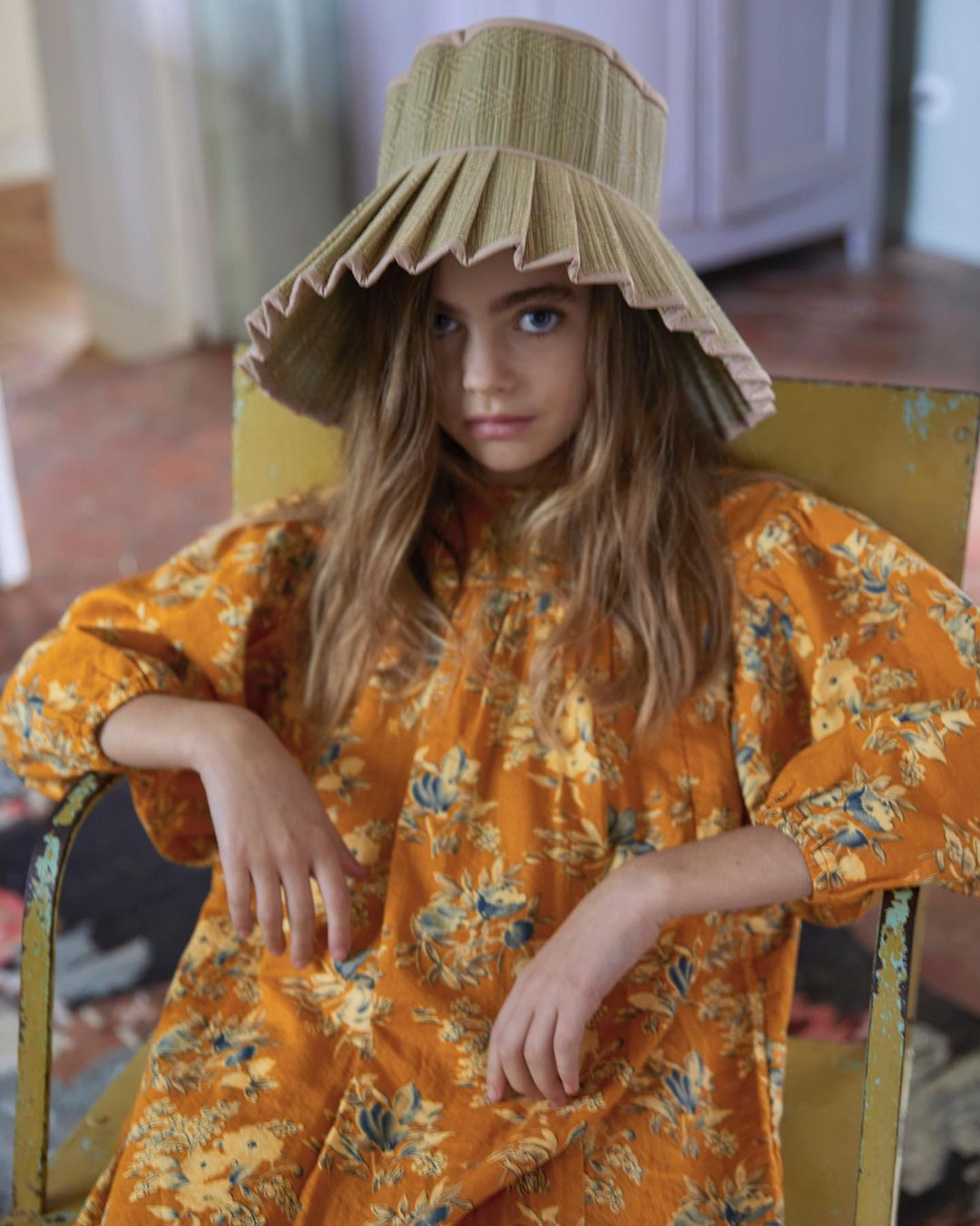 A young girl wearing a floral dress and a large sun hat sits indoors in a chair, looking directly at the camera with a neutral expression.