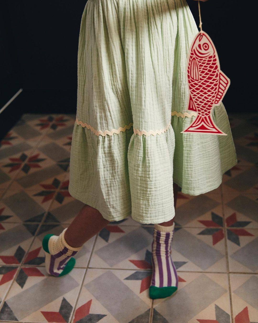 Child's lower half wearing striped socks and a light green dress, holding a red and white fish-shaped decoration indoors.