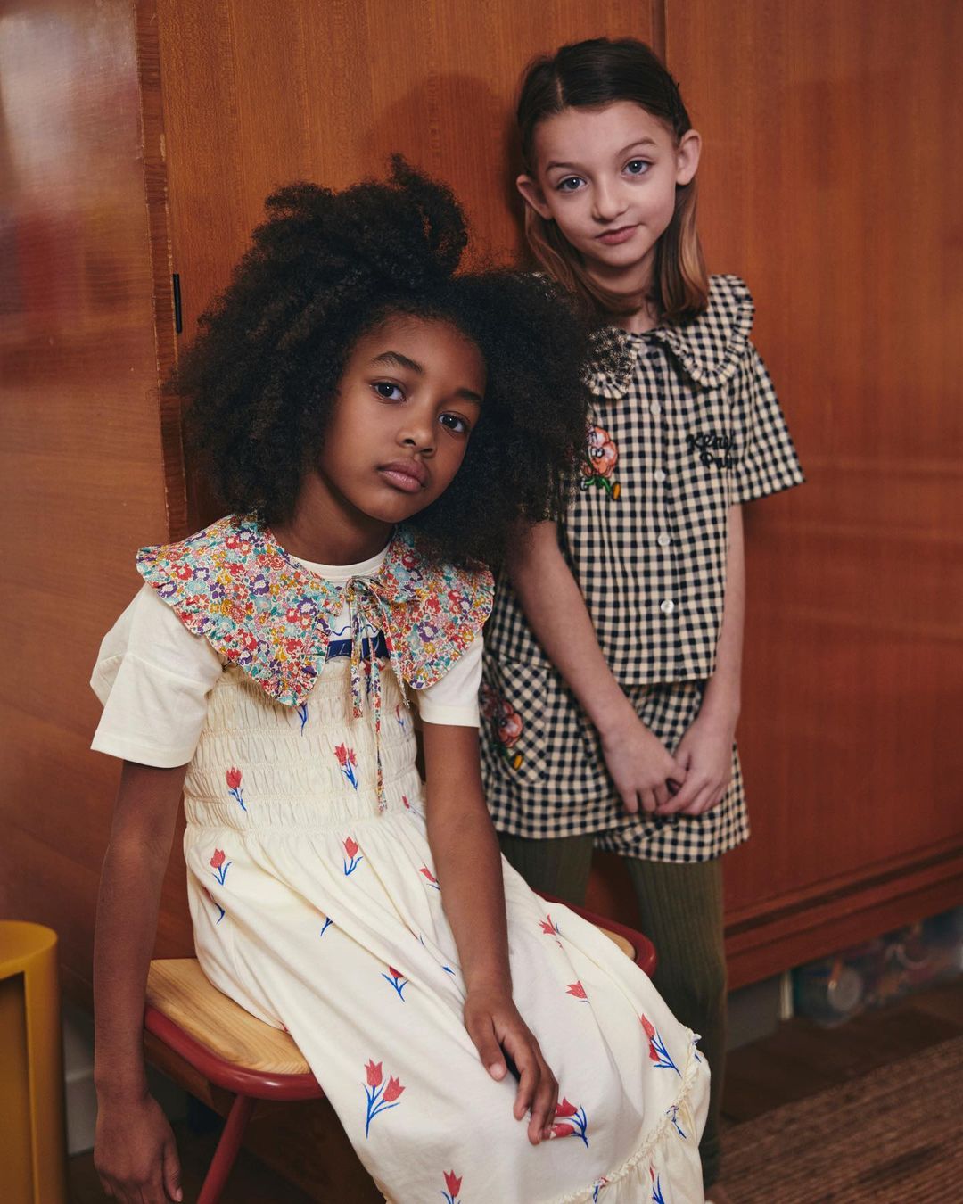 Two young girls pose indoors, one seated in a floral-collared dress, and the other standing in a black-and-white checkered outfit.