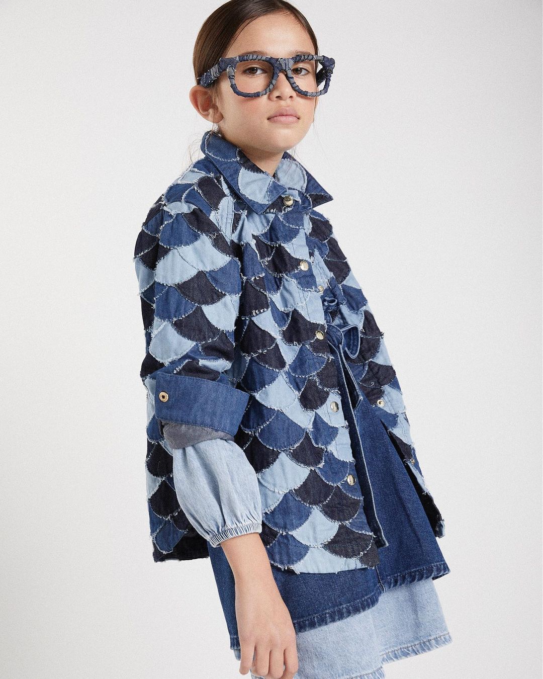 Girl in denim jacket and glasses poses in studio with neutral background. Jacket is pieced with varied blue denim. 