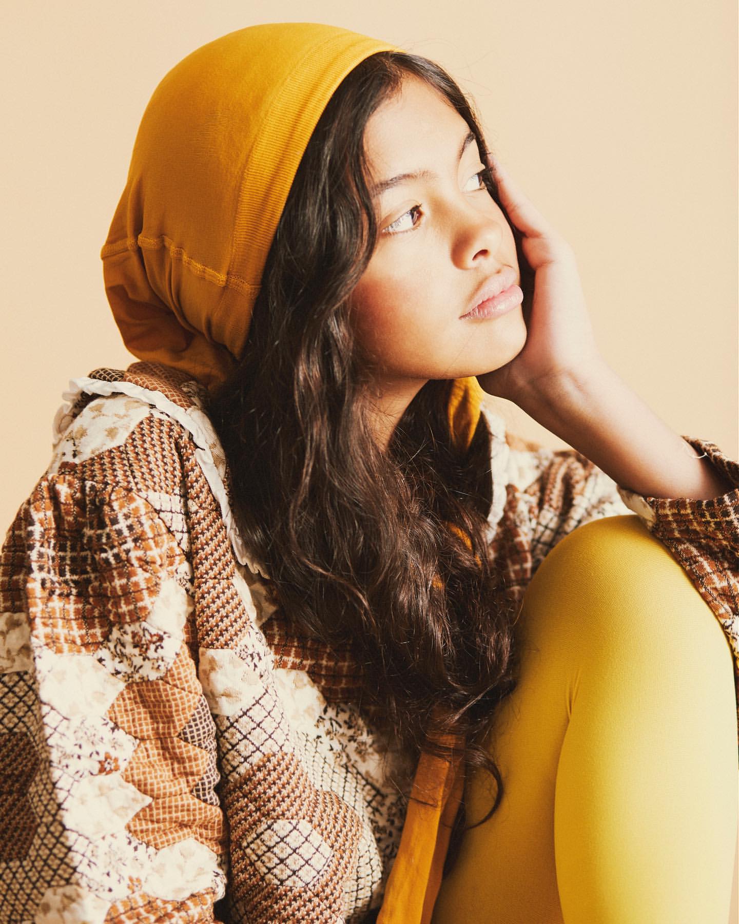 Portrait of a young woman with long brown hair wearing a yellow headscarf and patterned jacket, looking off to the side in a studio setting.