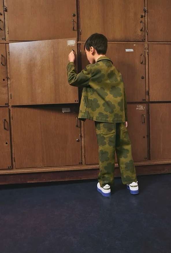 Back view of a child in a green floral print suit writing on a wooden locker. The setting is an indoor space with a blue floor.