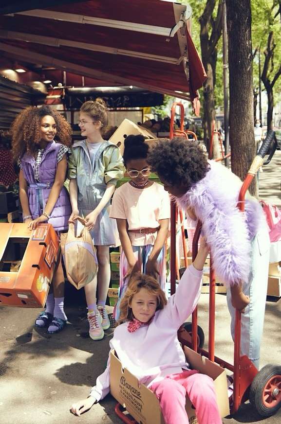 Group of five children in retro clothing outdoors with banana boxes and a dolly in an urban setting with an awning and trees.