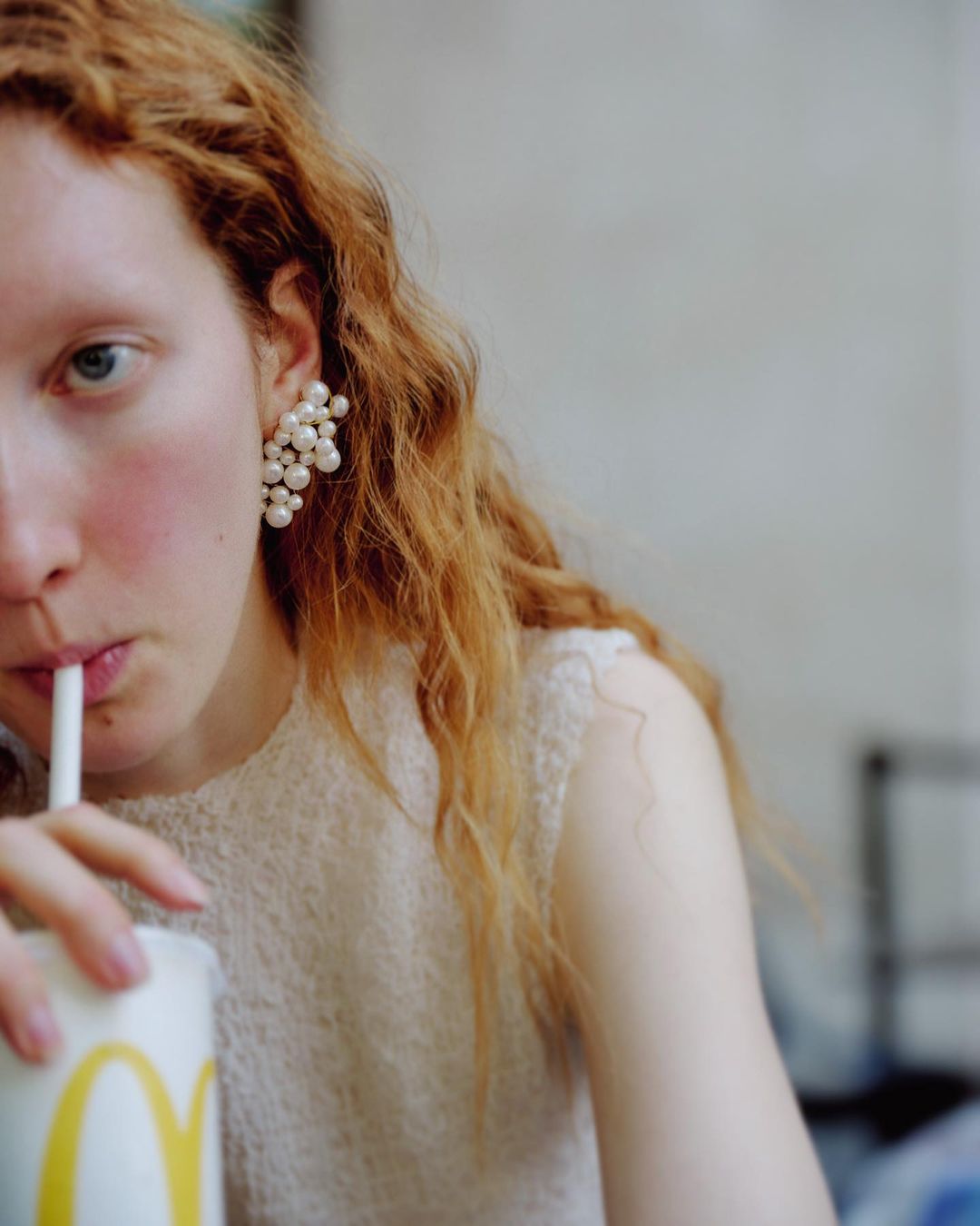 Close-up of a woman with curly red hair drinking from a McDonald's cup, wearing pearl earrings in a brightly lit interior.