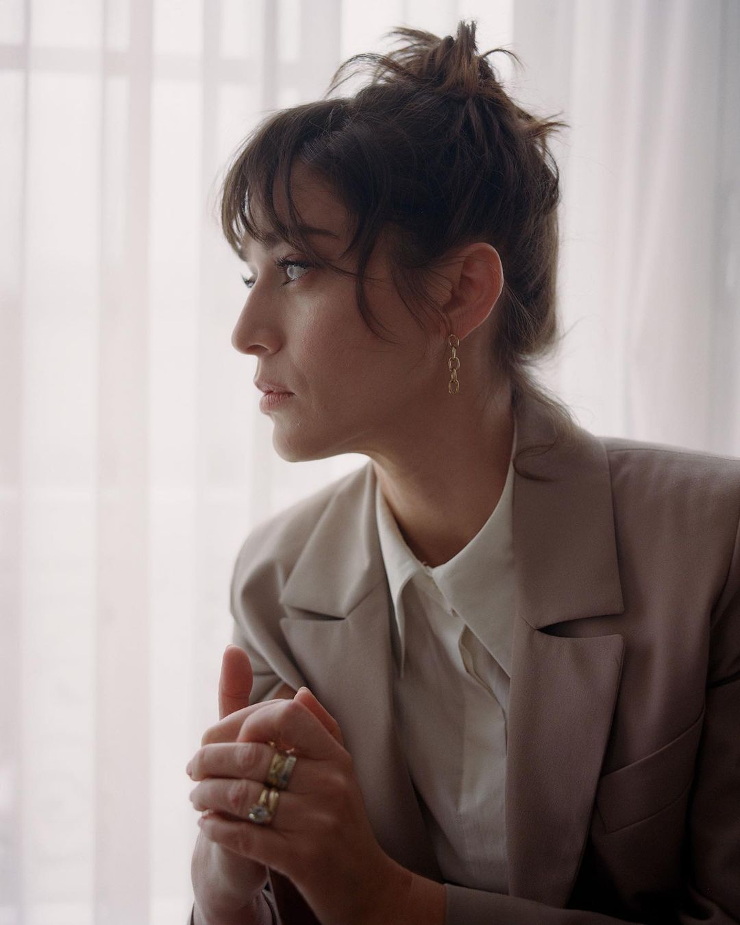 Portrait of a brunette woman with an updo, wearing a beige blazer and blouse, with hands clasped, in front of window light.