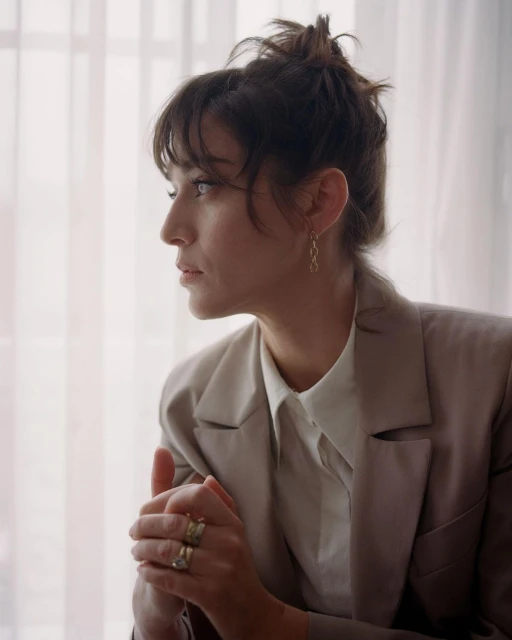 Portrait of a brunette woman with an updo, wearing a beige blazer and blouse, with hands clasped, in front of window light.