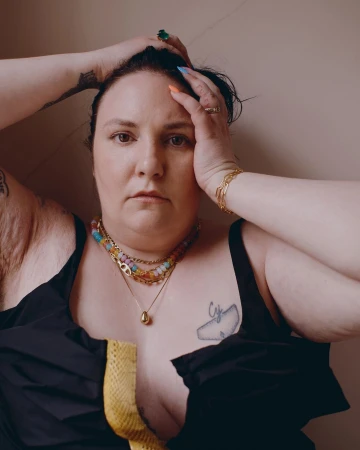 Portrait of a woman wearing layered beaded necklaces and a black top against a plain wall by Charlotte Hadden