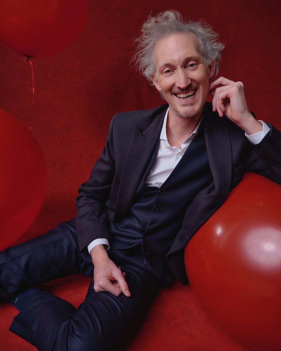 Smiling man in a dark suit poses against a vibrant red backdrop dotted with red balloons, exuding a cheerful and stylish vibe.