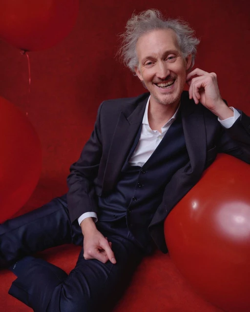 Smiling man in a dark suit poses against a vibrant red backdrop dotted with red balloons, exuding a cheerful and stylish vibe.