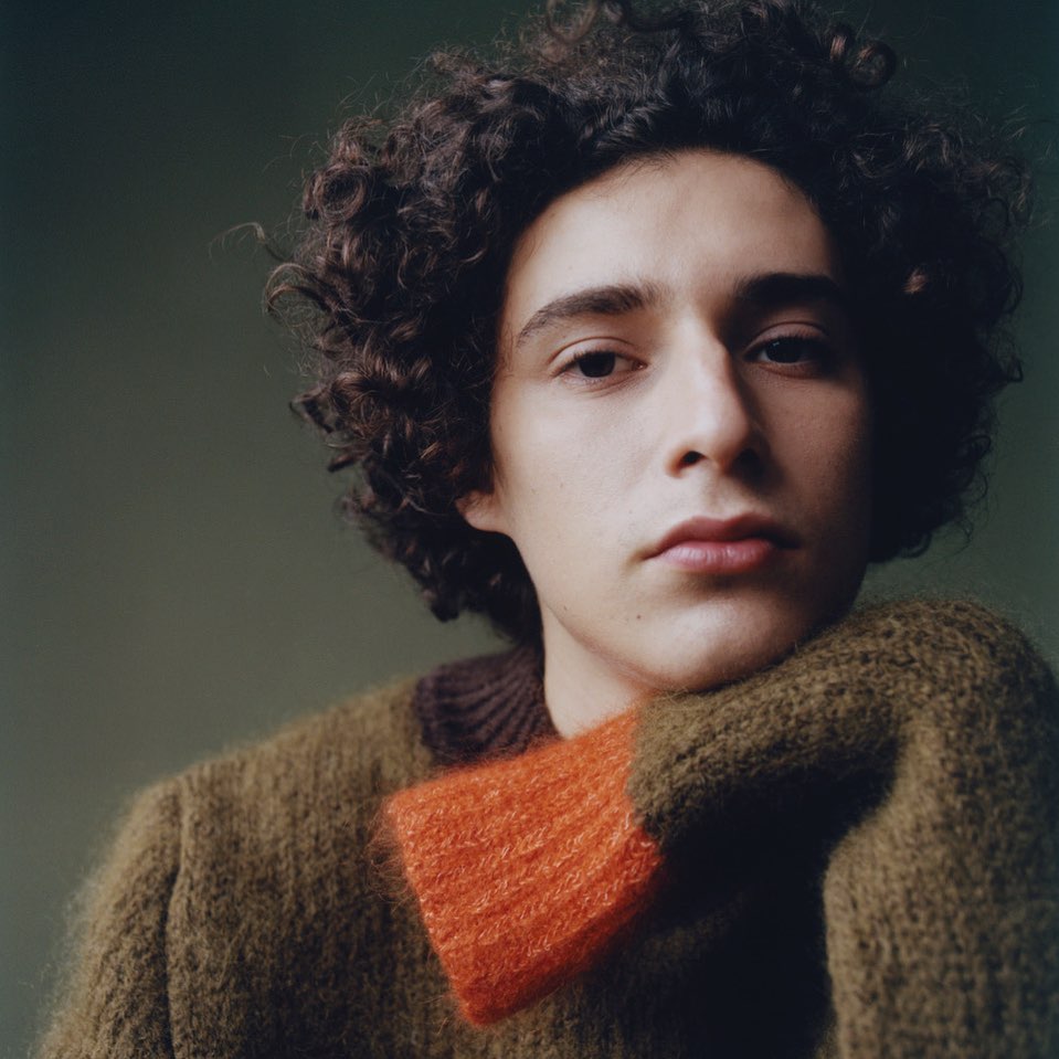 Close-up portrait of a young man with dark curly hair wearing an olive green and orange sweater against a dark background.