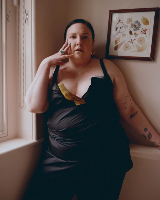 Portrait of an adult female with dark hair posing indoors. She wears a black top with gold detail. Flowers are framed on the wall.