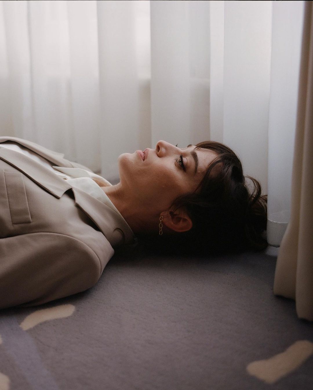 Brunette woman with fair skin lies on a grey patterned carpet in a beige suit, looking up towards white curtains in an indoor setting.