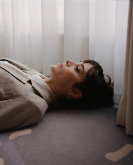 Brunette woman with fair skin lies on a grey patterned carpet in a beige suit, looking up towards white curtains in an indoor setting.