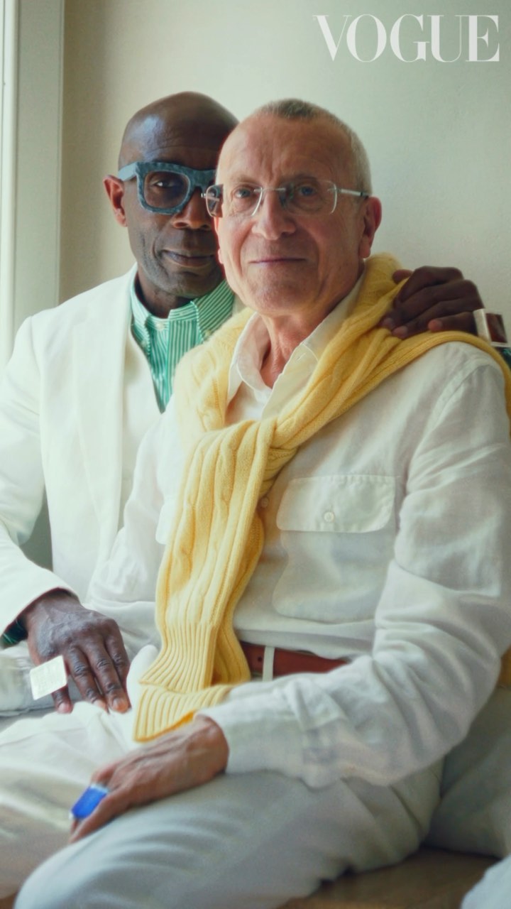 Portrait of two mature adult men in light colored clothing, one with a yellow sweater draped over his shoulders. Vogue logo visible.