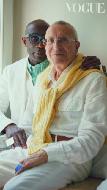 Portrait of two mature adult men in light colored clothing, one with a yellow sweater draped over his shoulders. Vogue logo visible.