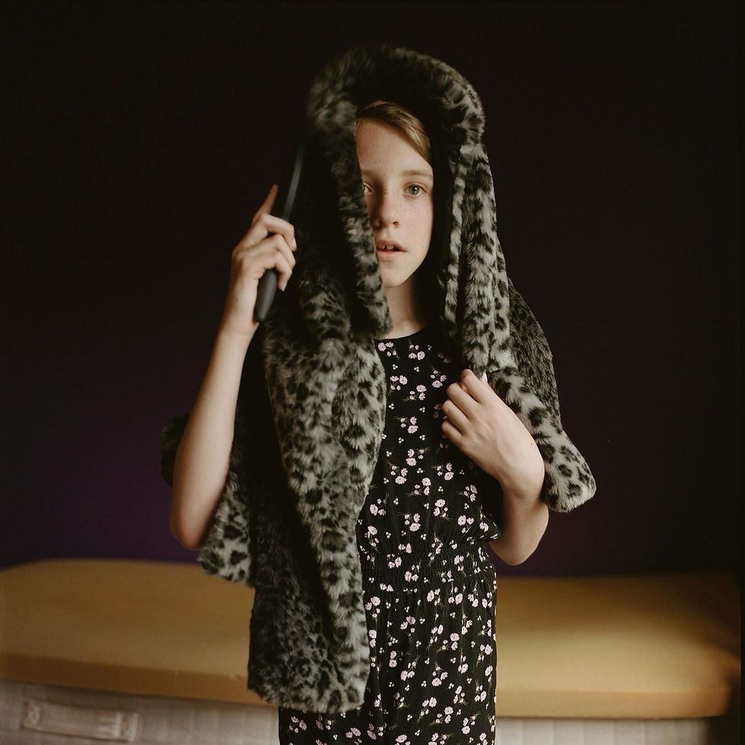 Young girl in a floral dress and faux fur leopard print hooded scarf poses in a studio with soft lighting, looking directly at the camera.