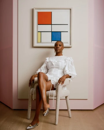 A bald Black woman wearing a white ruffled dress and metallic silver shoes seated on a modern textured chair.