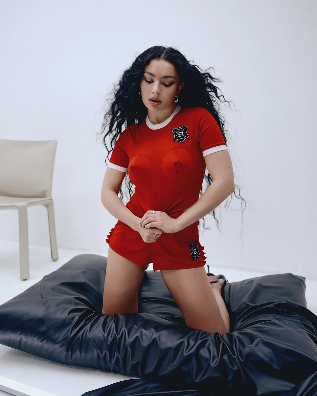 A woman with long dark curly hair kneels on a black satin cushion, wearing a red and white athletic-style outfit in a white studio.