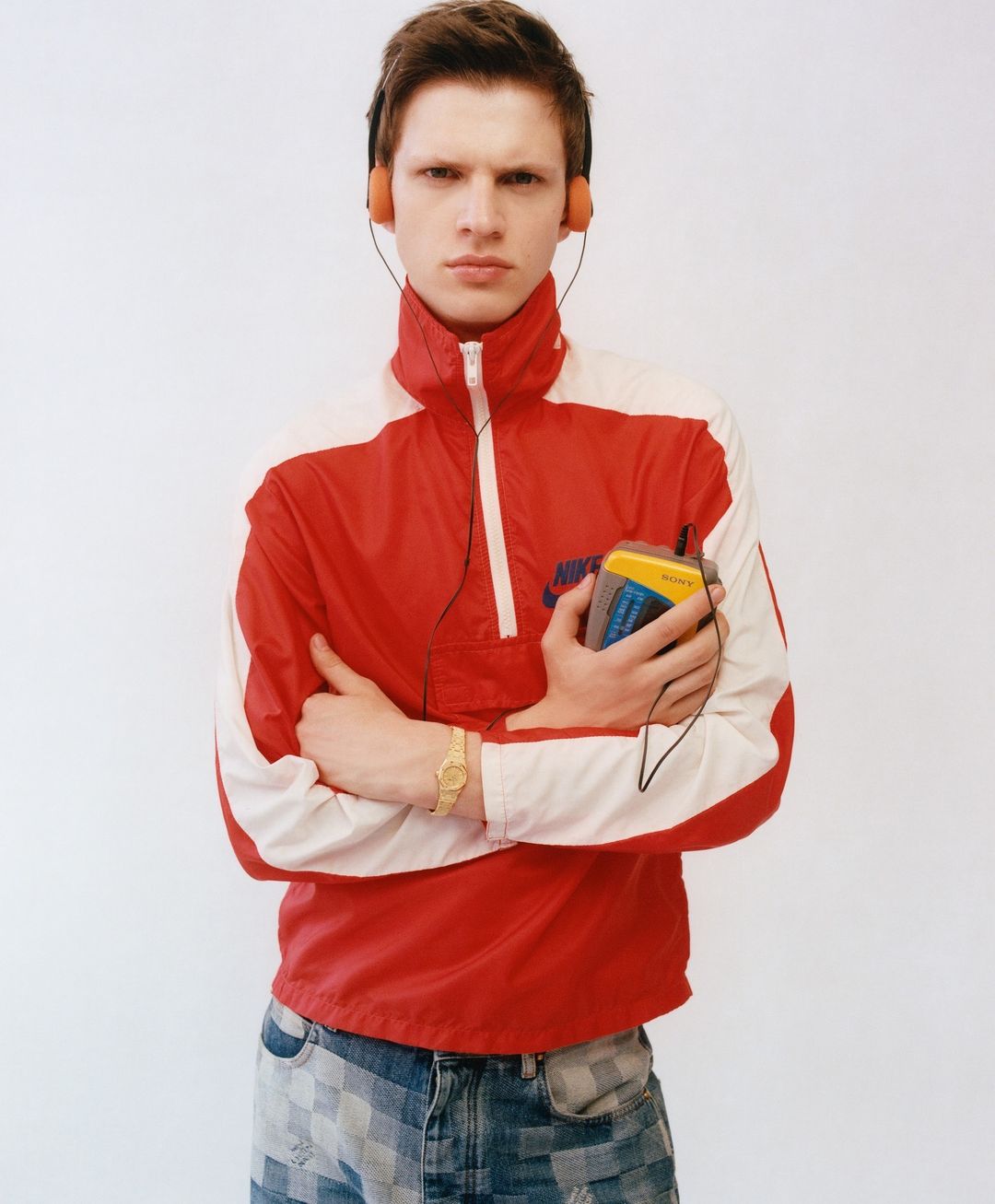 Man in red and white jacket holding retro Sony Walkman with orange headphones against a white wall backdrop, portrait style.
