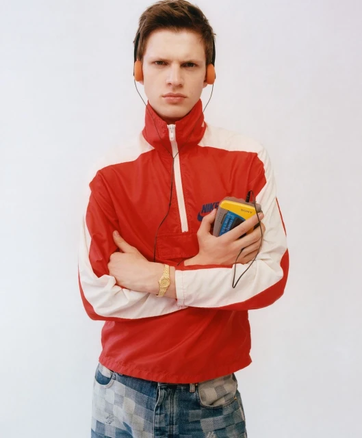 Man in red and white jacket holding retro Sony Walkman with orange headphones against a white wall backdrop, portrait style.