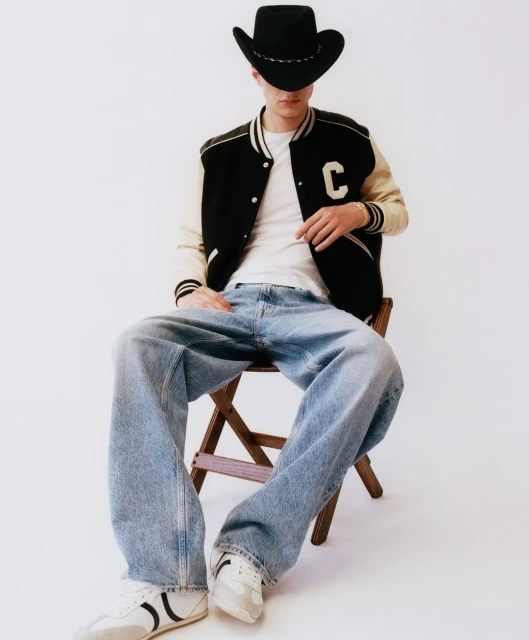 Man in college letterman jacket, light blue jeans, and black cowboy hat poses seated on wooden chair in a brightly lit studio.