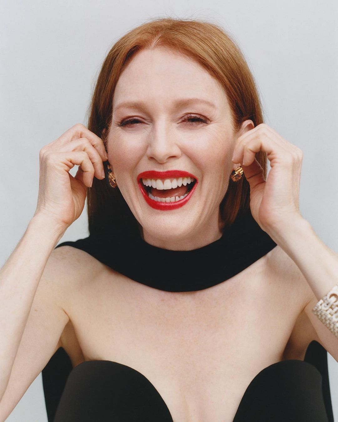 Smiling red-haired woman in a strapless black dress with a cape, accessorized with bracelet and earrings, poses against a plain background.