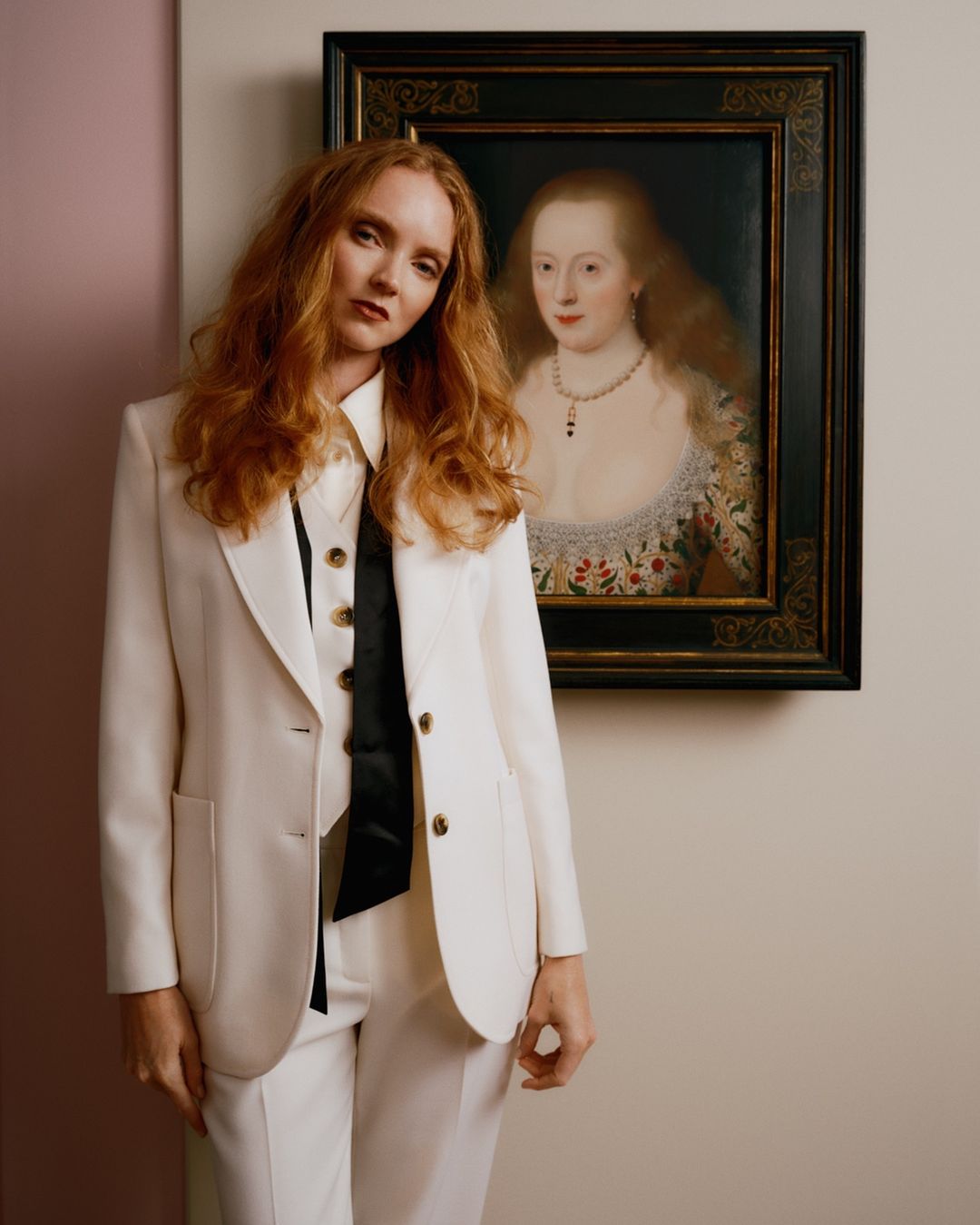 A red-haired woman in a white suit stands indoors next to a framed portrait of a woman in period attire, creating a vintage fashion aesthetic.