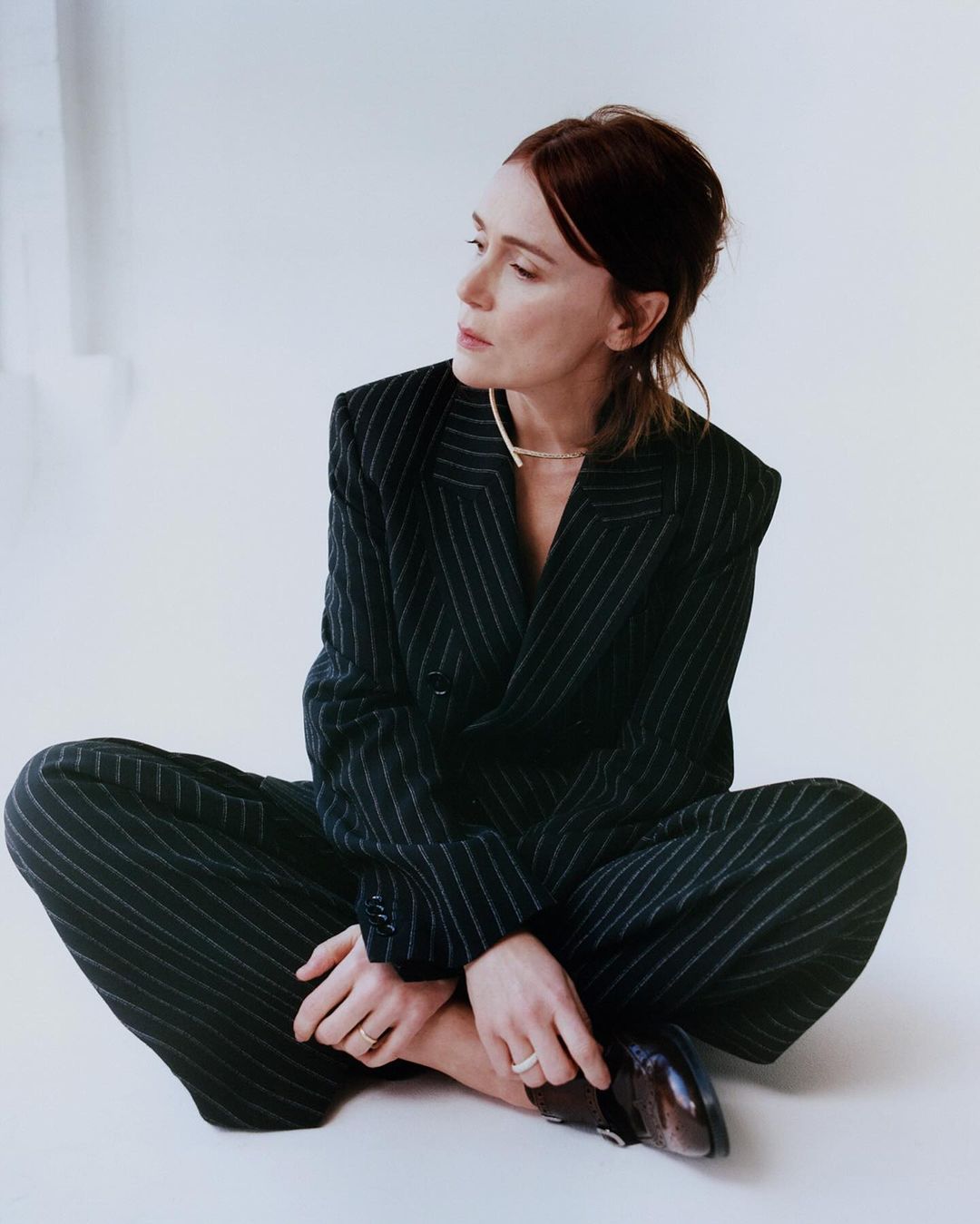 Stylish woman in a pinstripe suit sits cross-legged on a white surface, looking off to the side, showcasing fashion and modern professional attire.